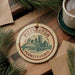 A circular ornament with the text "PITTSBURGH PENNSYLVANIA" and an illustration of a bridge and city skyline, hanging on a wooden surface.