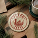 A circular ornament with the text "PLANO TEXAS U.S.A." printed on it, surrounded by pine branches and a cup of coffee on a wooden surface.