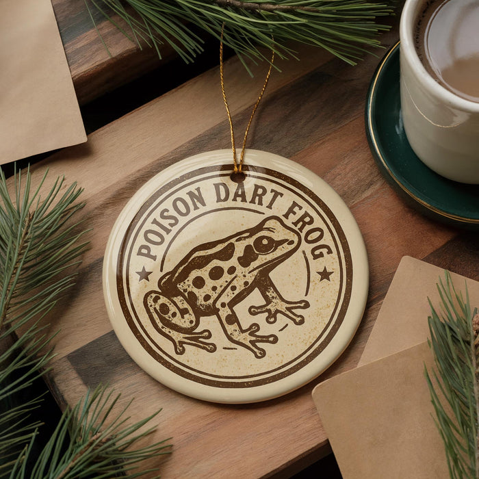 a round ornament with a brown and white design depicting a Poison Dart Frog, placed on a wooden surface alongside a cup of coffee and some pine branches.