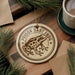 a round ornament with a brown and white design depicting a Poison Dart Frog, placed on a wooden surface alongside a cup of coffee and some pine branches.