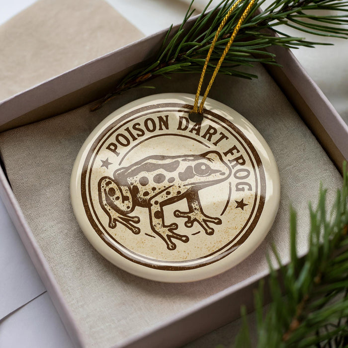 A round ornament with a brown and white design featuring a frog, placed inside a box with pine branches.