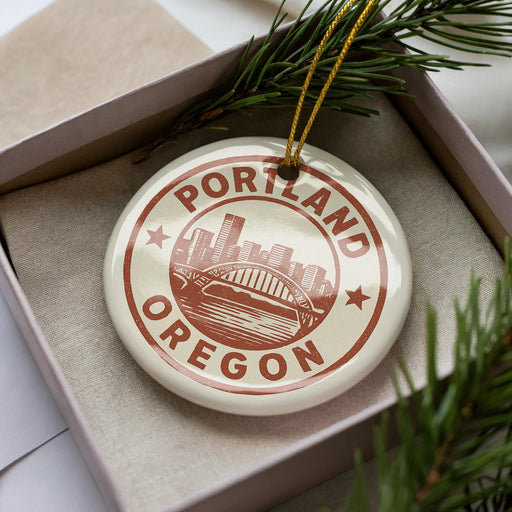 A decorative ornament in the shape of a city skyline, with the text "PORTLAND OREGON" prominently displayed, is placed in a box surrounded by pine branches.