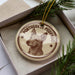 A circular ornament with a dog's head and the text "PORTUGUESE PODENGO" printed on it, placed inside a box with pine branches.