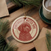 A round ornament featuring a red and white illustration of a Portuguese Water Dog, with the text "Portuguese Water Dog" printed on it, is displayed on a wooden surface alongside a cup of coffee and some pine branches.
