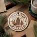 A circular ornament with the text "Prambanan Indonesia" and an illustration of a temple, placed on a wooden surface surrounded by pine branches and a cup of coffee.