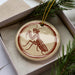 A circular ornament featuring a detailed illustration of a praying mantis, with a red and beige color scheme.