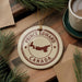 A circular ornament with the text "PRINCE EDWARD CANADA" and a map of Prince Edward Island, a region in Canada, is displayed on a wooden surface alongside a cup of coffee and some pine branches.