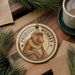 A round ornament featuring a monkey with the text "PROBOSCIIS MONKEY" on it, placed on a wooden surface next to a cup of coffee and some pine branches.