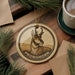 A round ornament featuring a close-up image of a pronghorn deer, with the word "PRONGHORN" written around the edge. The ornament is hanging from a string, and there is a cup of coffee and some pine branches in the background.
