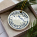 A white ornament with blue text and a map of the Bahamas, placed in a box with pine branches.