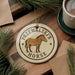 A round ornament featuring a horse is displayed on a wooden surface, surrounded by pine branches and a cup of coffee.