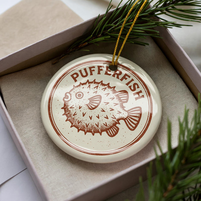 A white ornament with a brown design featuring a pufferfish is displayed in a box, surrounded by pine branches.