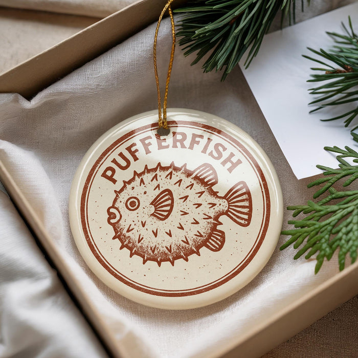 A round ornament with the text "PUFFERFISH" printed on it, featuring a pufferfish illustration, is displayed in a box with pine branches.