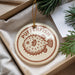 A round ornament with the text "PUFFERFISH" printed on it, featuring a pufferfish illustration, is displayed in a box with pine branches.