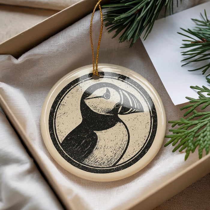 A decorative ornament featuring a black and white illustration of a bird, possibly a puffin, is displayed in a box with pine branches.