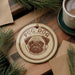 A round ornament featuring a pug dog design, with the text "PUG DOG" printed on it, is displayed on a wooden surface alongside a cup of coffee and some pine branches.