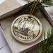 A decorative ornament in a box, featuring a circular design with a drawing of a building and the text "PECS HUNGARY" printed on it.