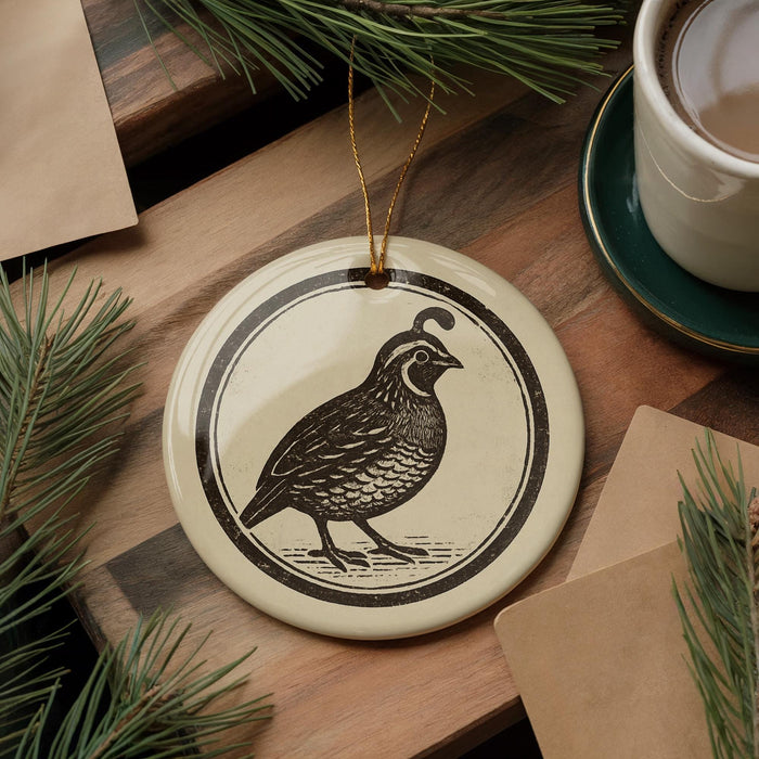 a ceramic ornament with a black and white illustration of a bird, likely a quail, on a wooden surface. The ornament is hanging from a string, and there is a cup of coffee and some pine branches in the background.