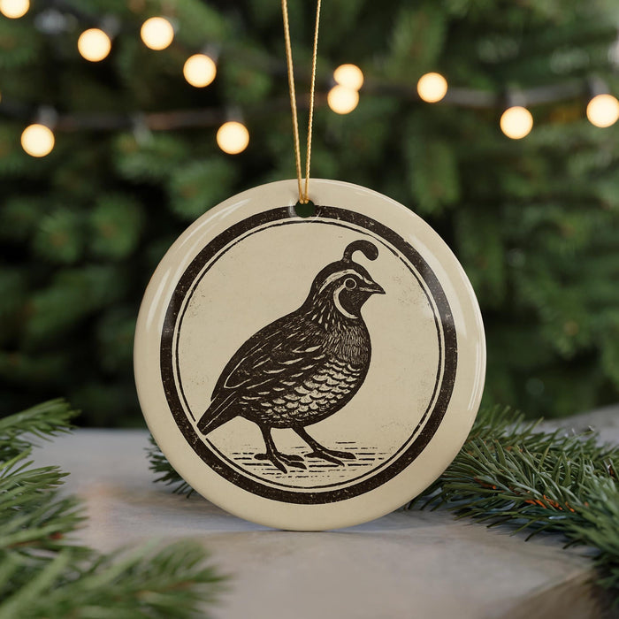 A round ceramic ornament featuring a black and white illustration of a quail bird is displayed in front of a blurred Christmas tree with twinkling lights.