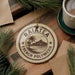 A circular ornament with the text "RAIATEA FRENCH POLYNESIA" printed on it, surrounded by pine branches and a cup of coffee on a wooden surface.