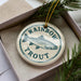 A white ceramic ornament with a blue and green design depicting a fish, specifically a rainbow trout, is displayed in a box with pine branches.