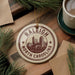 A decorative ornament featuring the Raleigh, North Carolina skyline, with pine branches and a cup of coffee nearby.