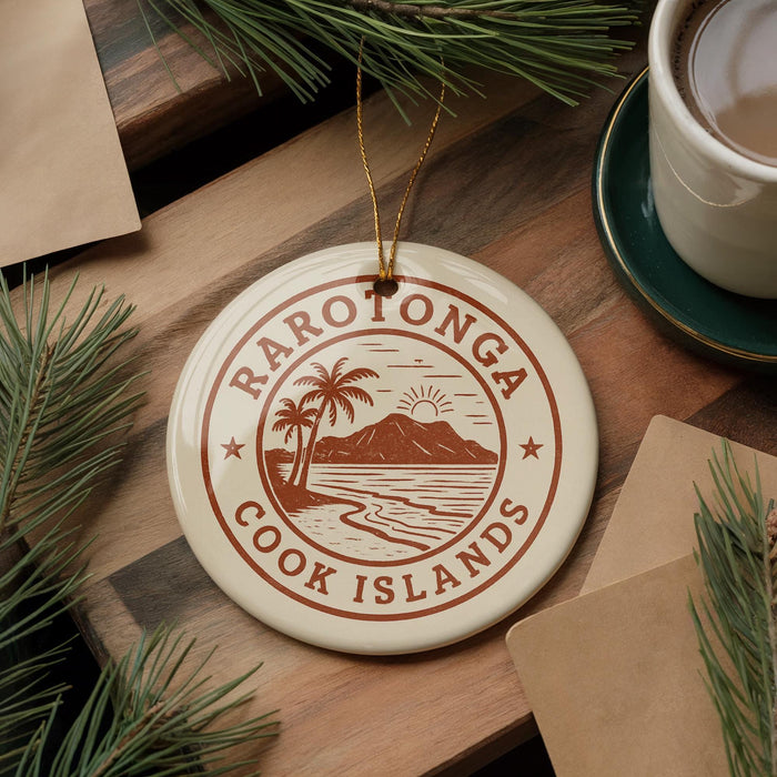 A circular ornament with the text "RAROTONGA COOK ISLANDS" and a palm tree illustration, placed on a wooden surface next to a cup of coffee and some pine branches.
