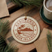 A circular ornament with the text "RAROTONGA COOK ISLANDS" and a palm tree illustration, placed on a wooden surface next to a cup of coffee and some pine branches.