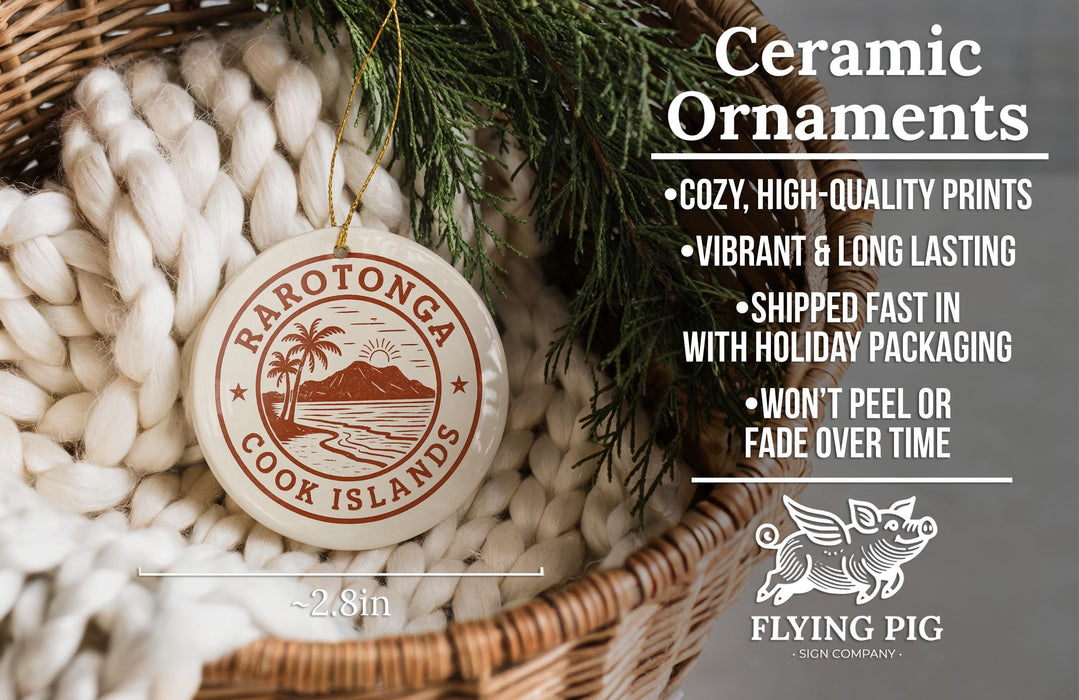 Ceramic ornaments, featuring a round ornament with a palm tree and a mountain, hanging in a basket with a pine branch.