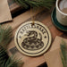 A round ornament with a snake design and the text "Rattlesnake" is displayed on a wooden surface, surrounded by pine branches and a cup of coffee.