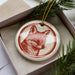 A ceramic ornament in the shape of a fox's head, with a red and white color scheme, is displayed in a box surrounded by pine branches.