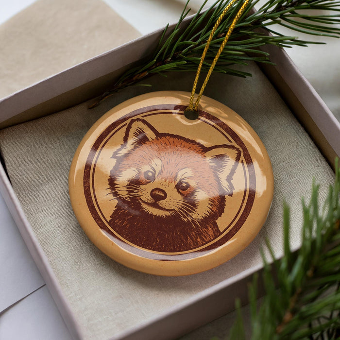 A ceramic ornament in the shape of a red panda, with a brown and beige color scheme, is displayed in a box surrounded by pine branches.