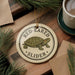 A round ornament featuring a turtle with the text "RED-EARED SLIDER" on it, placed on a wooden surface with pine branches and a cup of coffee nearby.
