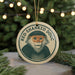 A round ornament featuring a monkey with a green and white pattern, hanging from a string against a blurred background of Christmas lights and greenery.