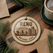 A circular ornament with the text "RENO NEVADA * USA" printed on it, surrounded by pine branches and a cup of coffee on a wooden surface.