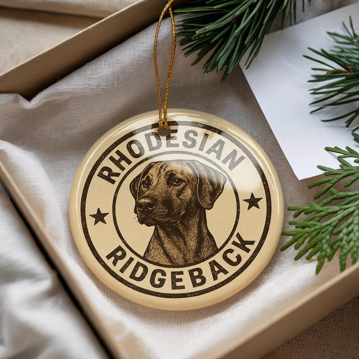 A round ornament with a dog image and the text "RHODESIAN RIDGEBACK" printed on it, placed in a box with pine branches.