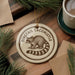A round ornament featuring a raccoon on a wooden surface, with a cup of coffee and pine branches in the background.