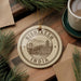 A decorative ornament with the text "RISHKESH INDIA" and an illustration of a bridge is displayed on a wooden surface, surrounded by pine branches and a cup of coffee.