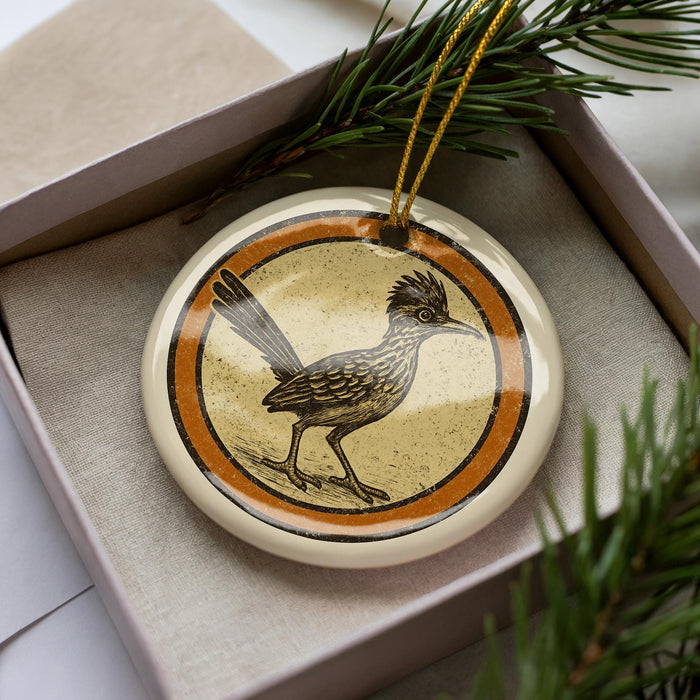 A decorative ornament featuring a bird, likely a roadrunner, is displayed in a box surrounded by pine branches.