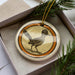 A decorative ornament featuring a bird, likely a roadrunner, is displayed in a box surrounded by pine branches.