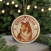 A round ornament featuring a collie dog, with the text "ROUGH COLLIE" printed on it, is hanging from a string in front of a Christmas tree.