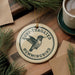 A round ornament with the text "RUBY-THROATED HUMMINGBIRD" and an illustration of a hummingbird is displayed on a wooden surface, accompanied by pine branches and a cup of coffee.
