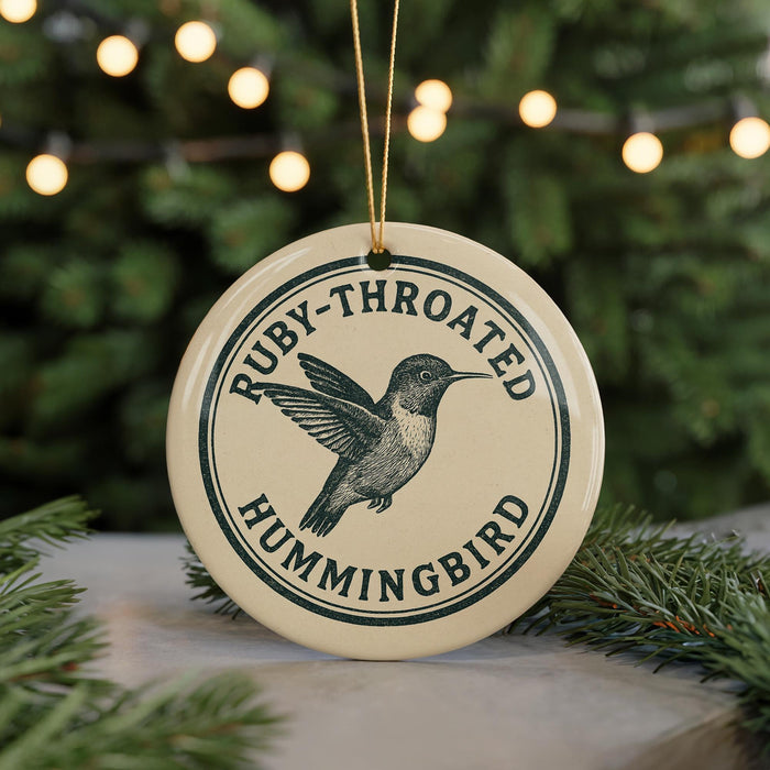 A round ornament with the text "Ruby-throated Hummingbird" and a graphic of a hummingbird is displayed on a table with pine branches in the background.