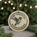 A round ornament with the text "Ruby-throated Hummingbird" and a graphic of a hummingbird is displayed on a table with pine branches in the background.