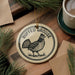 A round ornament featuring a pheasant, with the text "RUFFED GROUSE" printed on it, is displayed on a wooden surface alongside a cup of coffee and some pine branches.