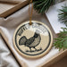 A decorative ornament featuring a bird, specifically a pheasant, with the text "RUFFED GOUSE" on it. The ornament is placed in a box with pine branches and a white paper.