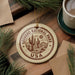 A circular ornament with the text "SAGUARO NATIONAL PARK USA" and an illustration of a cactus is displayed on a wooden surface, surrounded by pine branches and a cup of coffee.