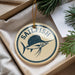 A round ornament with the word "SAILFISH" printed on it, featuring a graphic of a sailfish. The ornament is hanging from a string and is placed inside a box with pine branches visible.