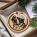 A round ornament featuring a dog's portrait is displayed in a box, surrounded by pine branches.