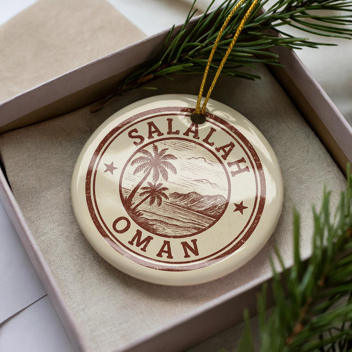 A circular ornament with the text "SALALAH OMAN" printed on it, surrounded by a palm tree and mountains, is displayed in a box with pine branches.